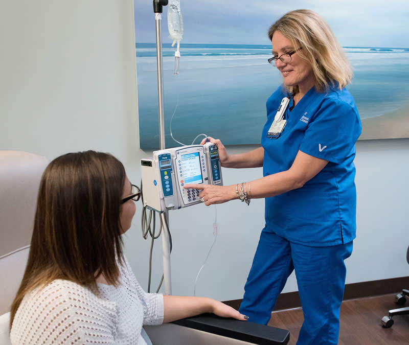 A female Baptist MD Anderson taps buttons on an infusion machine, talking with a female patient seated comfortably.
