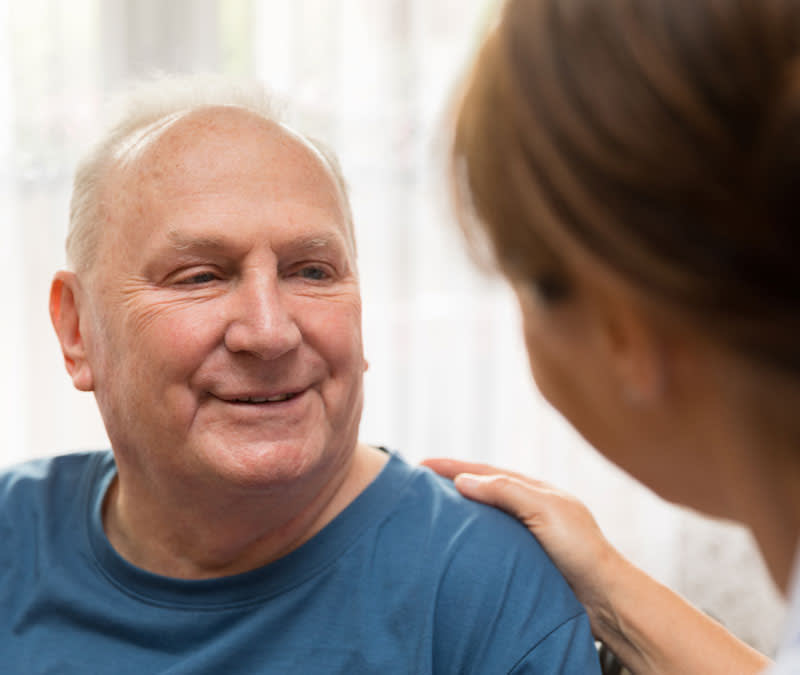 A senior man smiles at a support services professional, her hand resting gently on his shoulder.