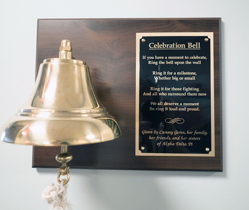 A golden bell with a plaque that says, 'Celebration Bell: If you have a moment to celebrate, ring the bell upon the wall. Ring it for a milestone, whether big or small. Ring it for those fighting, and all who surround them now. We all deserve a moment, so ring it loud and proud.'