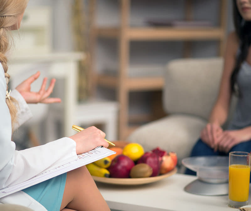 A female nutritionist sits with a patient, a bowl of healthy fruits and vegetables between them.