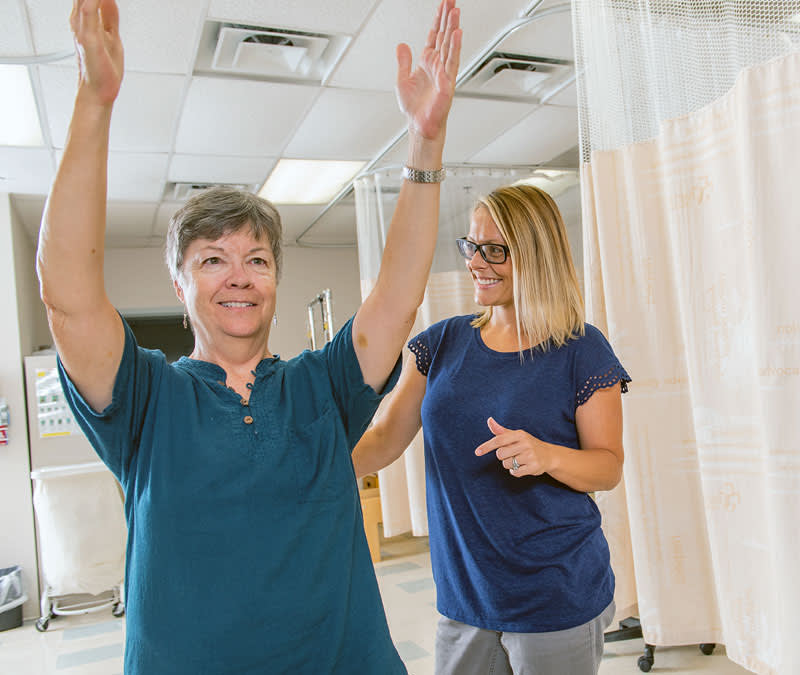 A female rehabilitation specialists works with a female patient, who stretches her arms over her head.