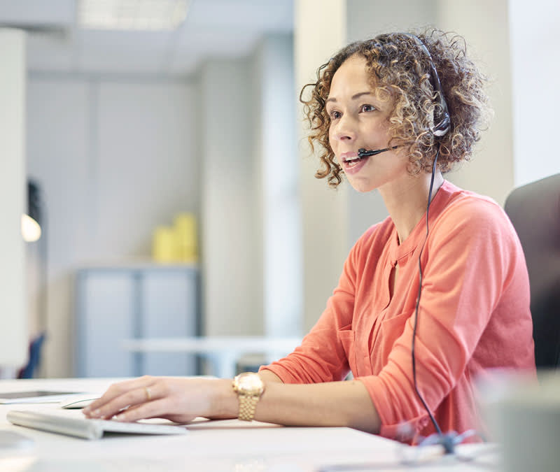 A curly-haired woman in bright coral sweater and wearing a headset assists a caller.