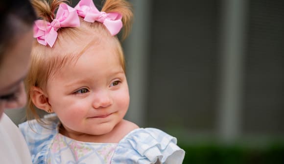 Rilynn, a pediatric patient with patent ductus arteriosus (PDA), smiles wearing a frilly dress with bows in her hair, being held my mom.