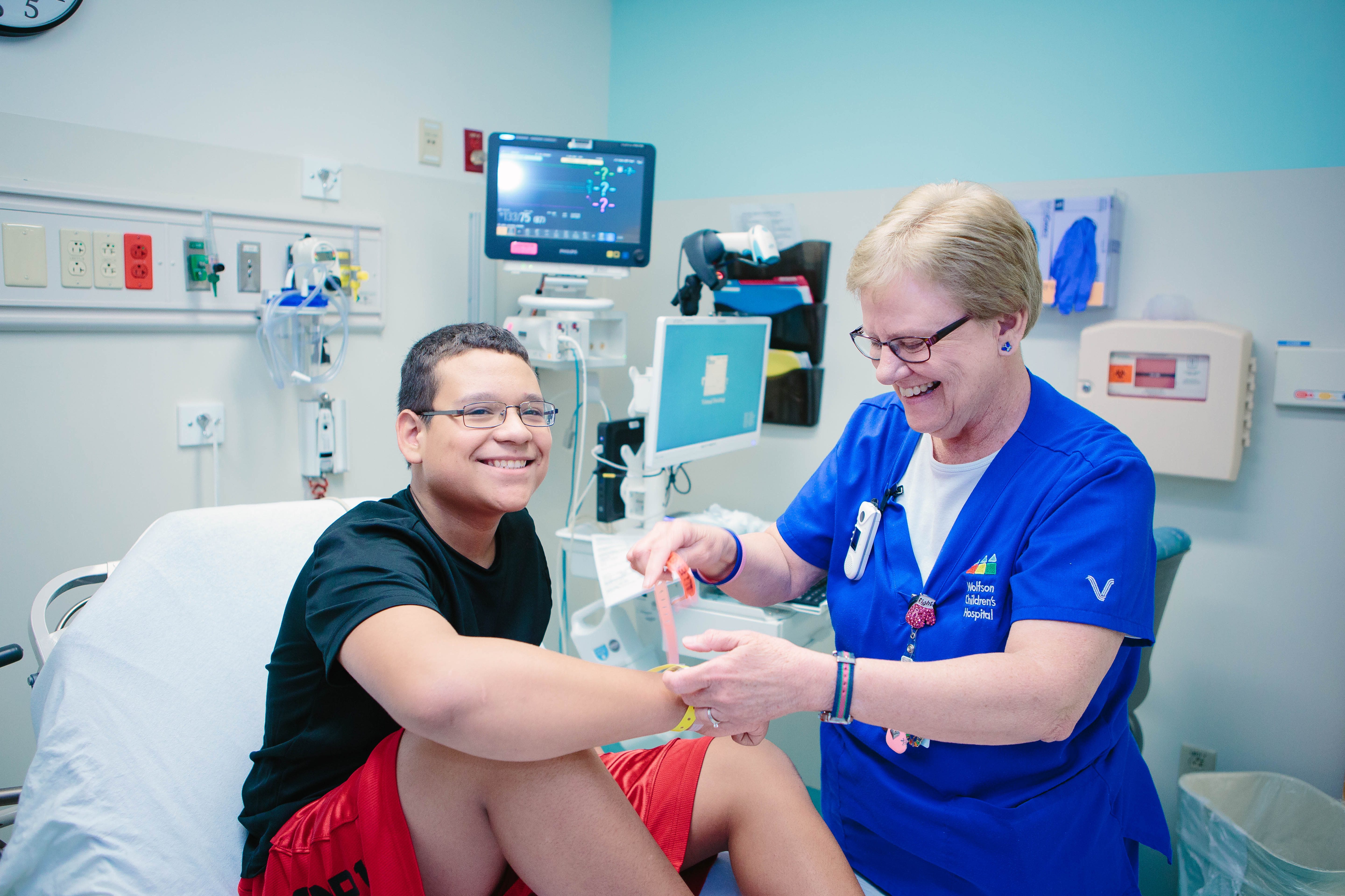 smiling teenage boy and patient at Wolfson Children's receives care. smiling female care team member attaches hospital bracelet to teen's wrist