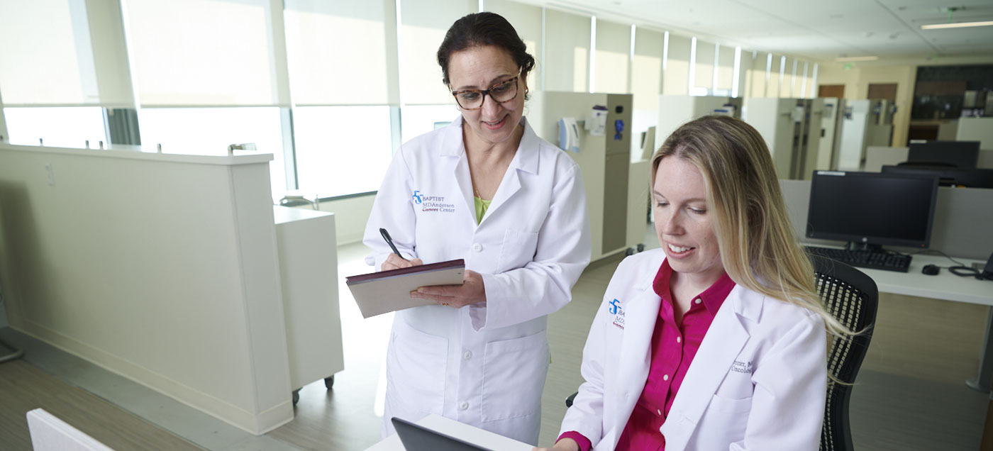 A female physician stands with a pen and pad of paper talks with another female physician at a computer looking through clinical research.