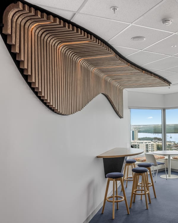 Custom Zintra Sticks installation in the Anindilyakwa Land Council Offices. The image shows a curved, wave-like ceiling feature made of wooden slats with integrated lighting, extending along a white wall above a high-top table and stools. A window with a cityscape view is visible in the background.