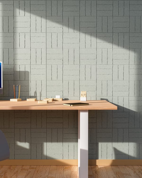 A modern workspace featuring a textured Organic Etch Panel Stacked wall in a soft blue-green hue. Sunlight casts dynamic shadows, highlighting the intricate etched patterns. A minimalist wooden desk with a height-adjustable white base adds functionality to the warm, contemporary setting.