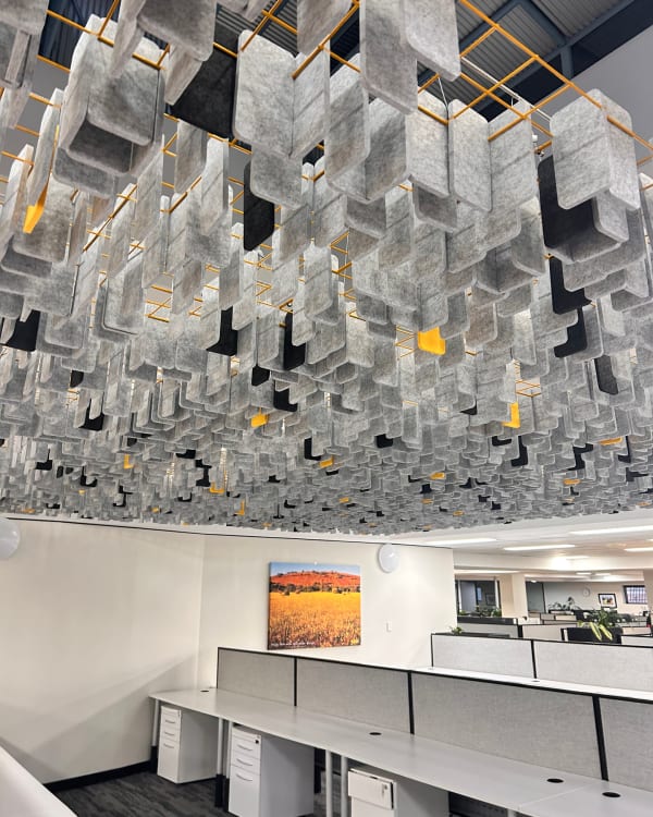 An open-plan corporate office features a modern acoustic ceiling installation using Zintra Mesh Baffles in a Random pattern. The grey, black, and yellow acoustic felt baffles are suspended from an orange grid frame over office cubicles, showcasing an innovative commercial ceiling design that balances sound absorption with vibrant architectural aesthetics.