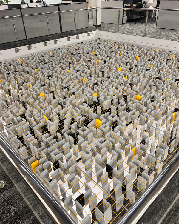 An aerial, top-down view of an office void featuring a large-scale installation of Zintra Mesh Baffles in a Random pattern. Suspended within the open atrium space the complex yellow wire grid organizes a dense layout of grey, black, and pops of yellow felt acoustic baffles, demonstrating an innovative approach to acoustic treatment in multi-level commercial interiors.