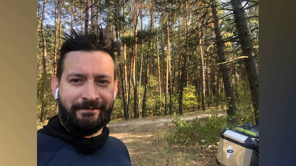 A man wearing a blue shirt is standing in front of a forest.