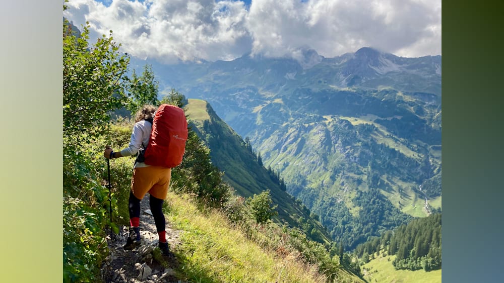 A woman with a red back pack hiking up a mountain with a clear blue sky in the background.