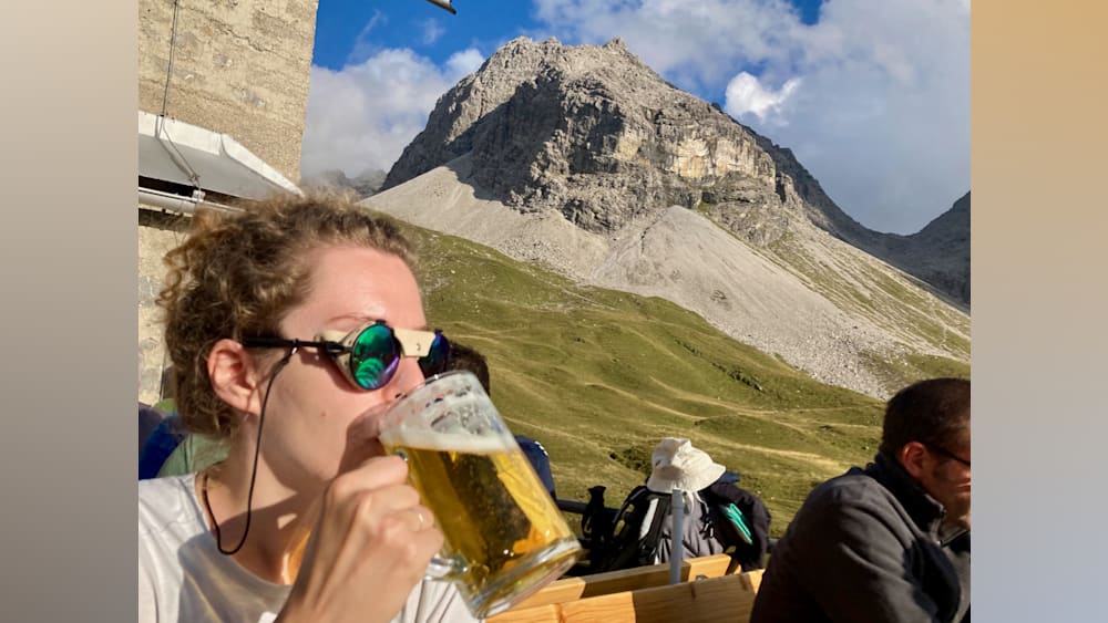 A woman wearing glasses drinks beer in front of a mountain.