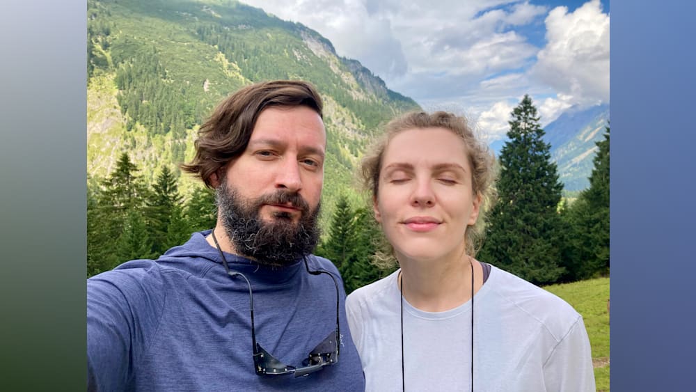 A man and a woman standing together in front of a forest wearing sunglasses.