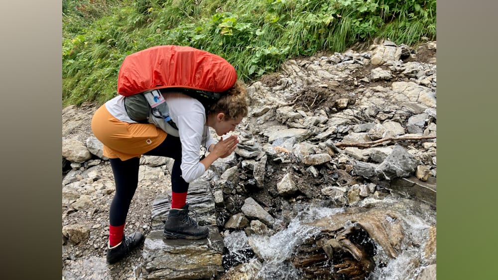 A woman is standing over a stream and taking a drink from her water bottle.