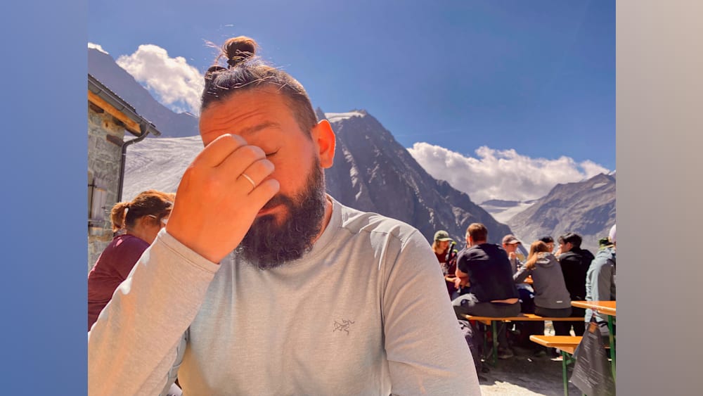 A bearded man with a long-sleeved shirt and a hand on his forehead sits at a table in the outdoors.