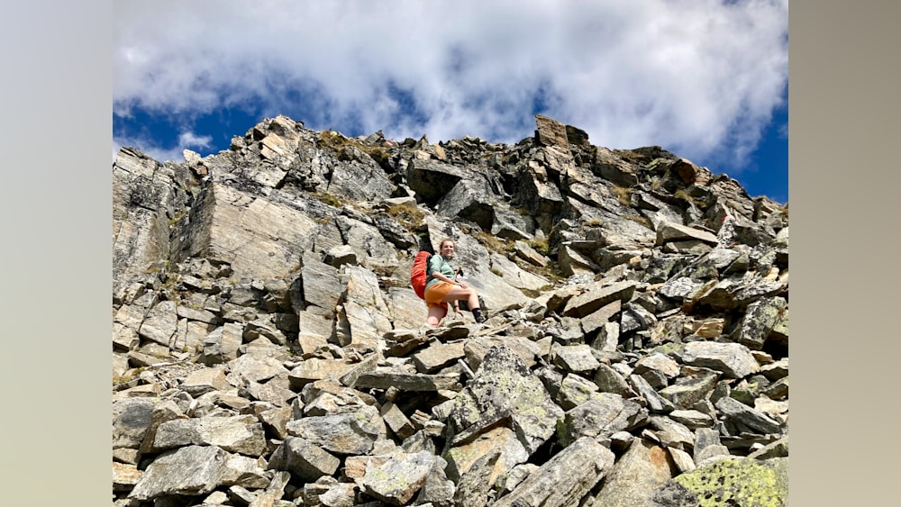 A woman in a red backpack is posing for a photo on a rocky mountain top.