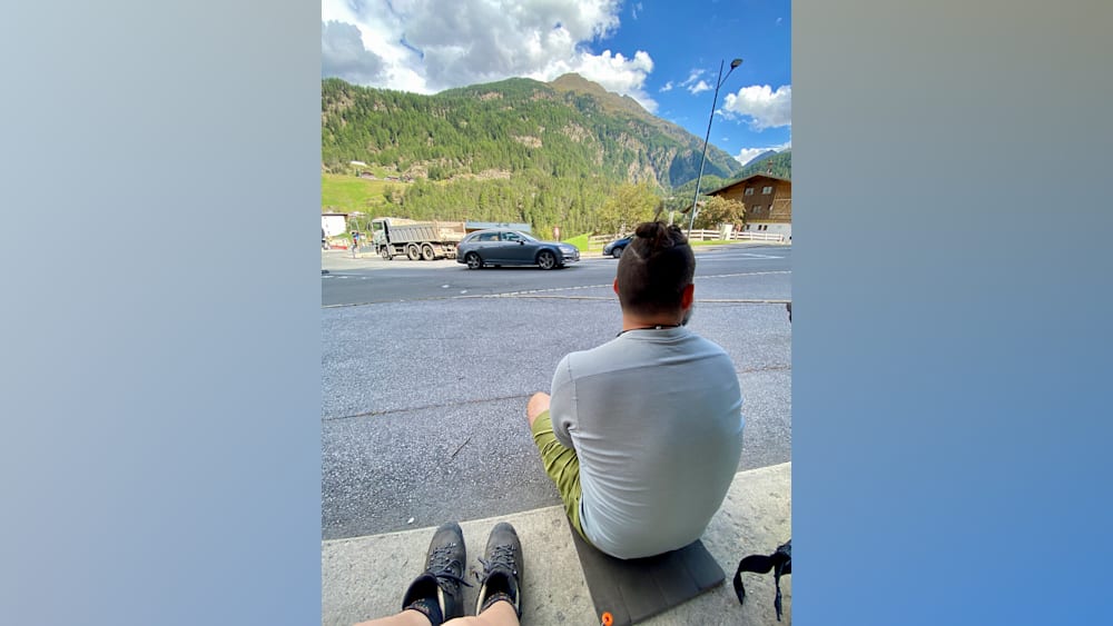 A man with a ponytail sits on the curb watching traffic pass by.