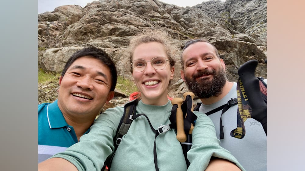 Three men and a woman are smiling for the camera in front of a rocky outcropping, with the woman wearing a backpack.