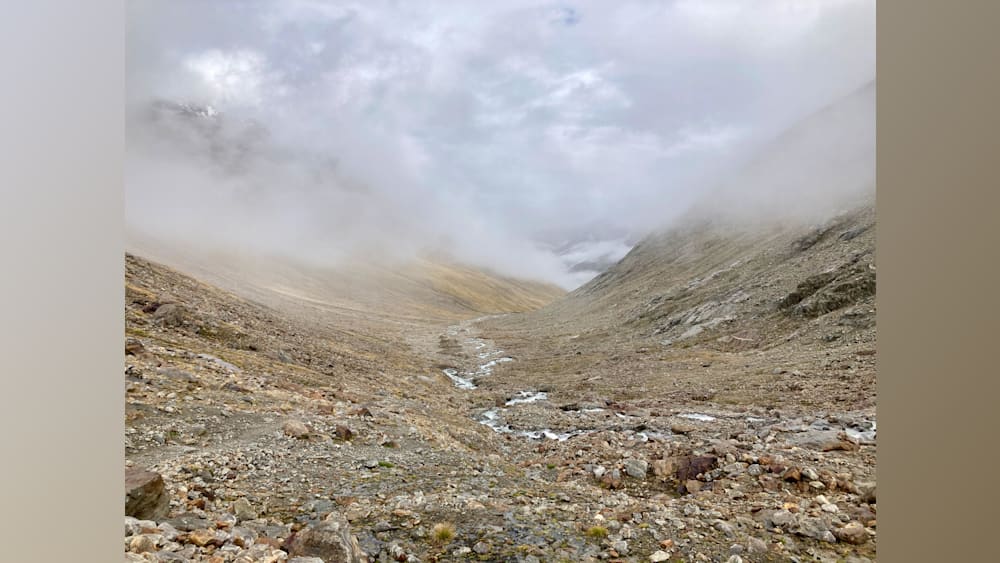 A foggy, rocky path winds through a valley with a stream running through it.