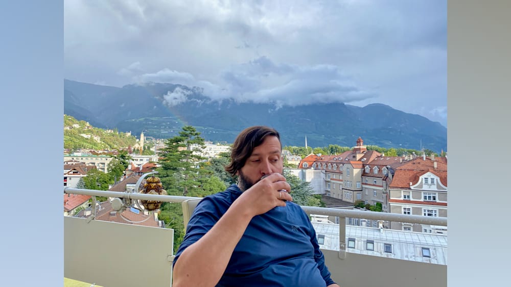 A man in a blue shirt is sitting outside and drinking from a glass.
