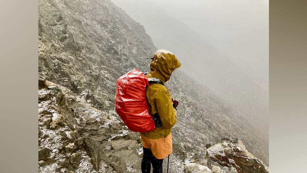 A man in a yellow jacket looks out over a snowy mountain range from the top of a hill.