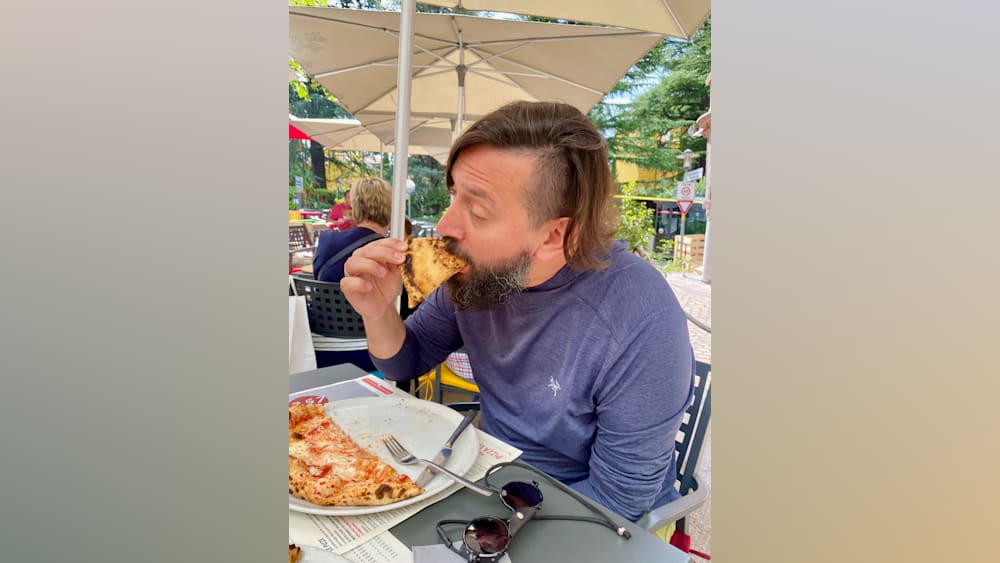 A man with a beard eating pizza in a blue shirt.