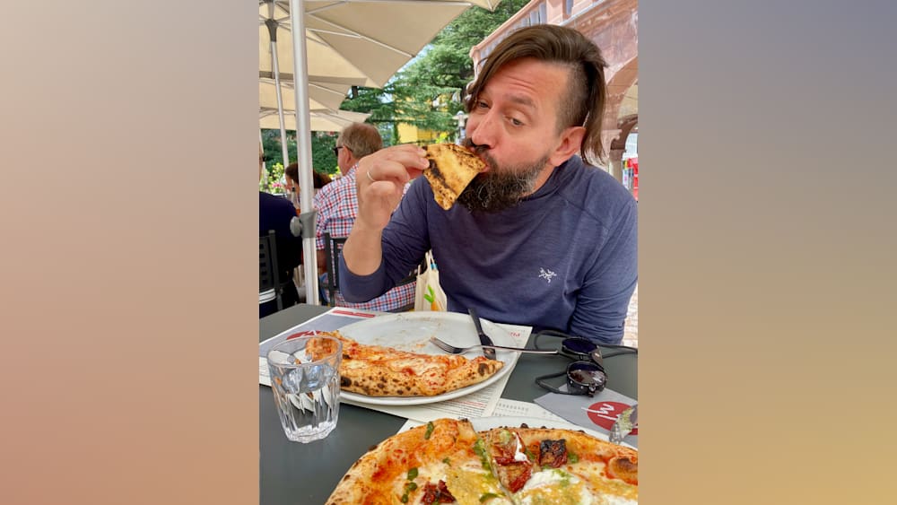 A man eating a slice of pizza at a table outside with sunglasses on top of the table.