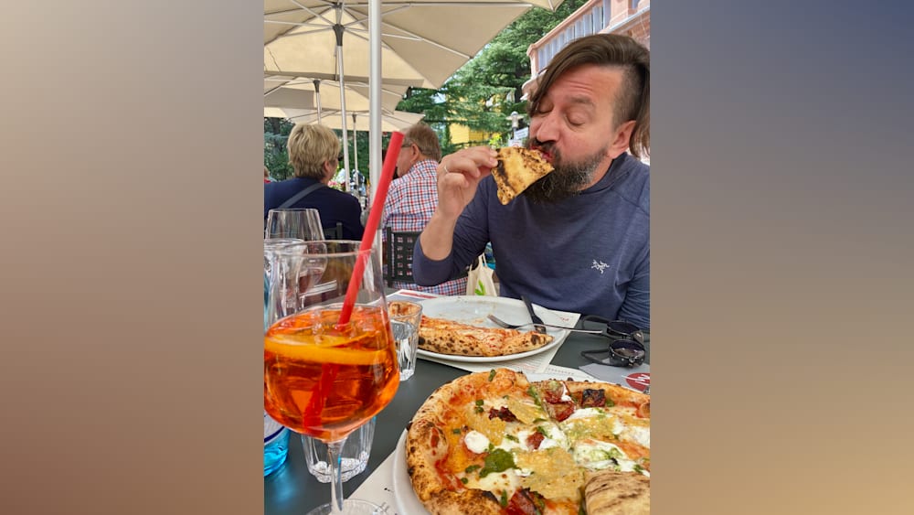 A man with a beard eating pizza and a drink with a straw.