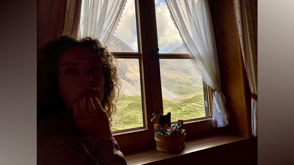A woman standing in front of a window with mountains in the background.