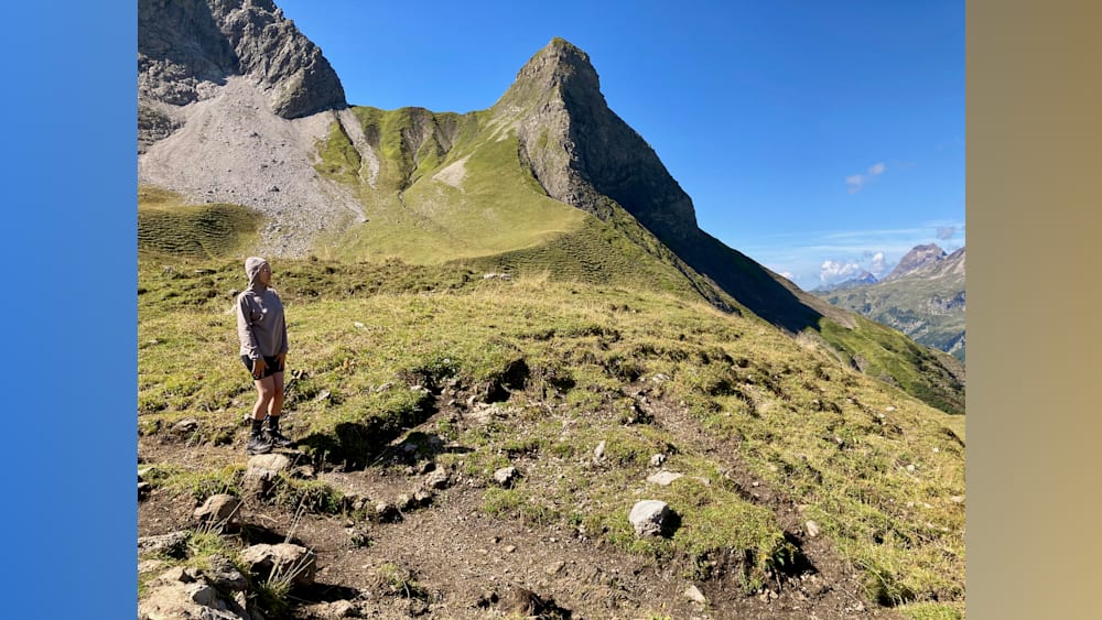 A woman stands at the top of a grassy hill with a large mountain in the background.