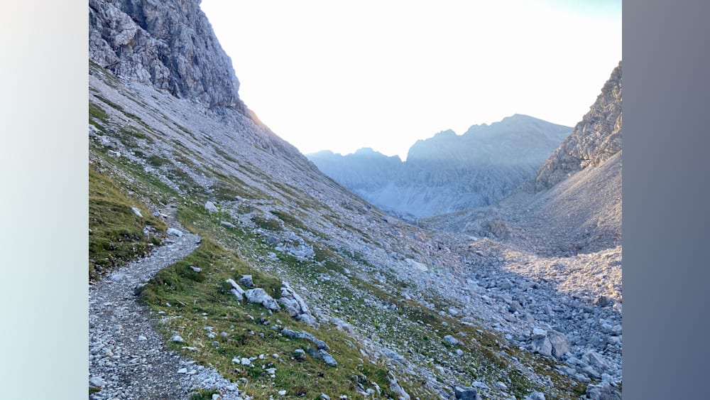 A path winds up the side of a mountain, through a grassy field, and up the side of a cliff.