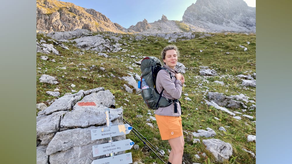 A woman wearing a grey and orange jacket stands on a mountain with a backpack on her shoulders.