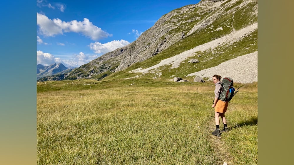 A woman with a backpack stands in a field looking at the mountains.