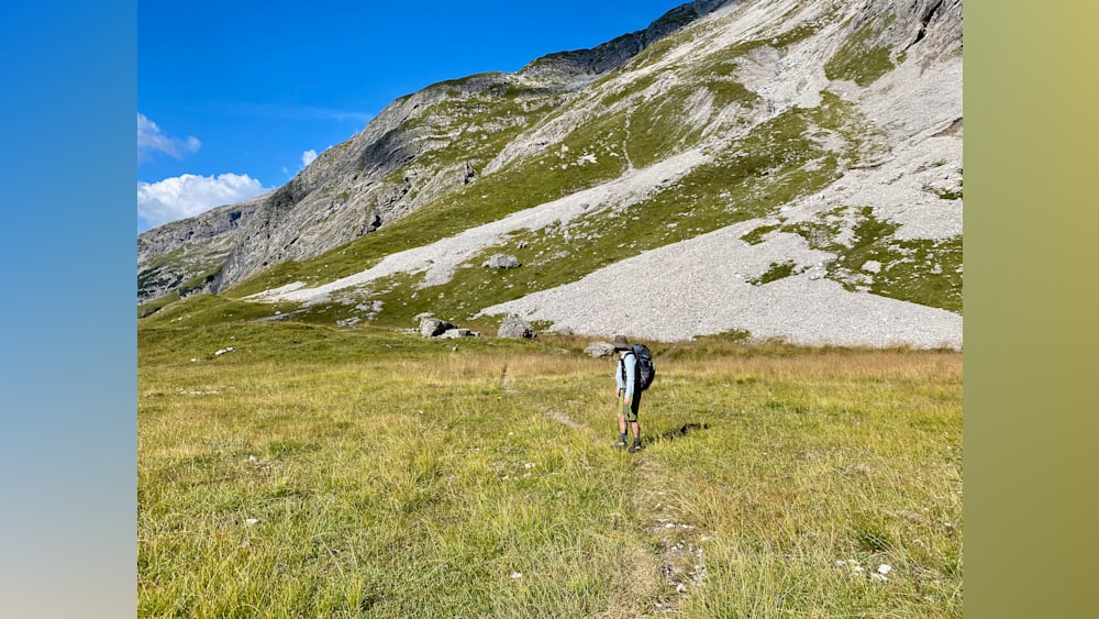A woman with a backpack stands in a field looking at a large hill.
