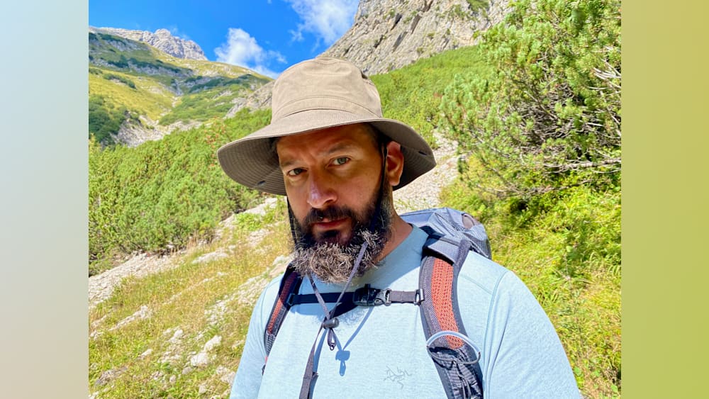 A man wearing a hat and backpack in front of a mountain.