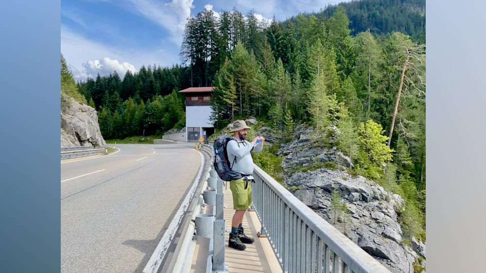 A man with a hat taking a picture on a bridge with his backpack on.