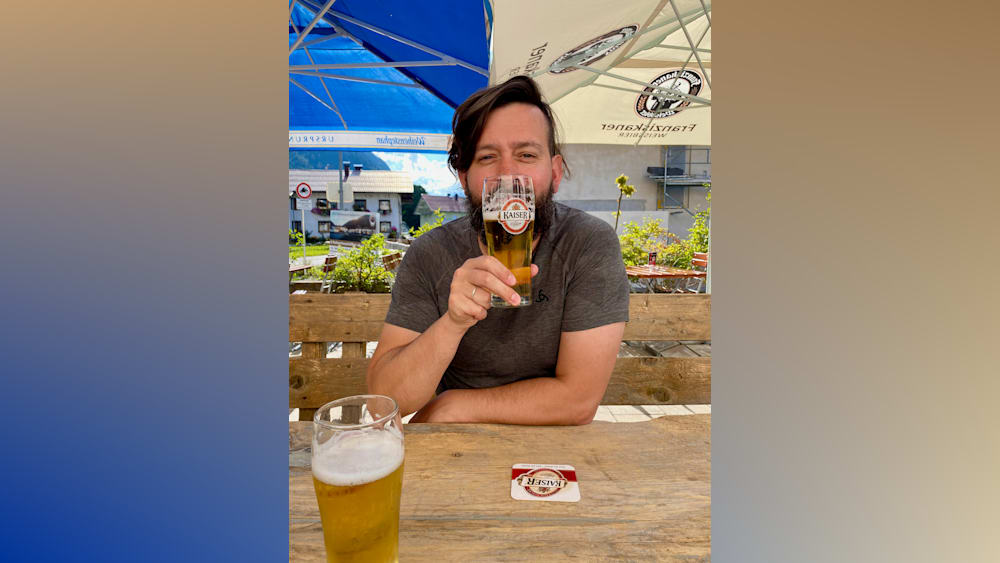 A man with a beard is holding a glass of beer up to his face and laughing.