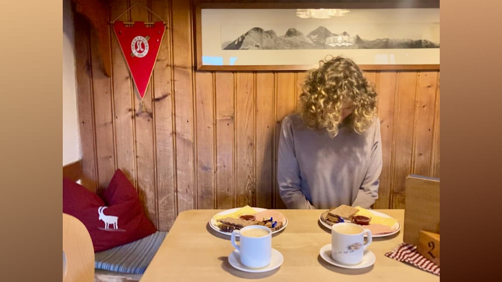A woman with curly hair sitting at a table with two plates of food and a cup of coffee in front of her.