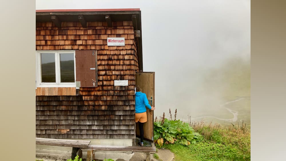 A woman in a blue jacket enters a cabin with wood shingles.