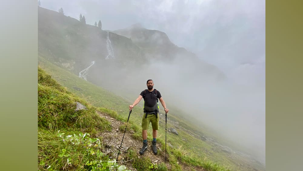A man standing on a mountain with his hiking poles up and a backpack on.