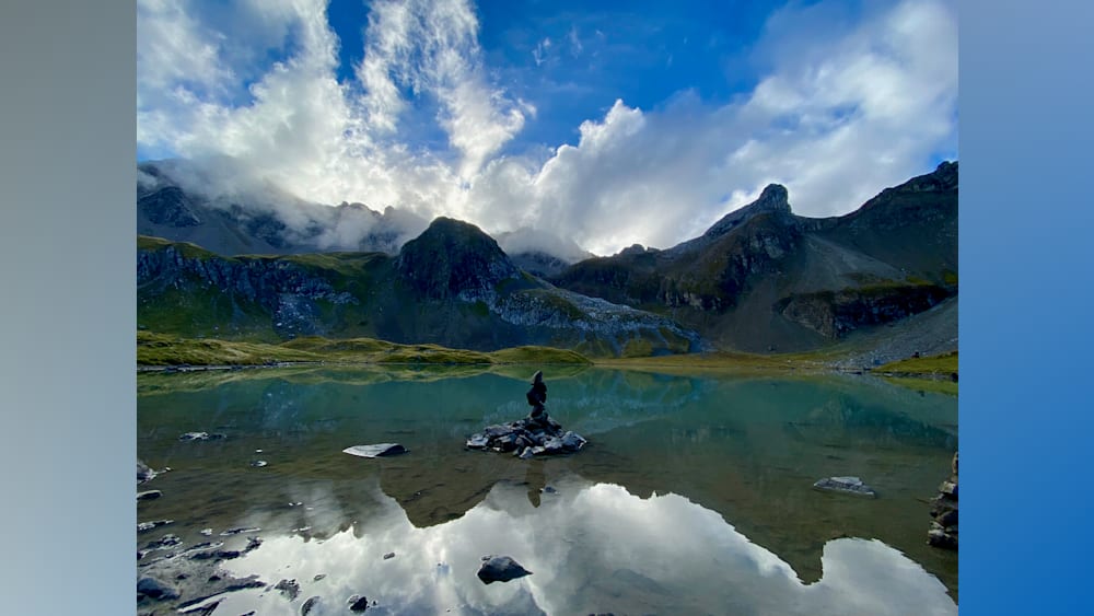 A man sits on a rock in front of a lake with clouds in the sky.