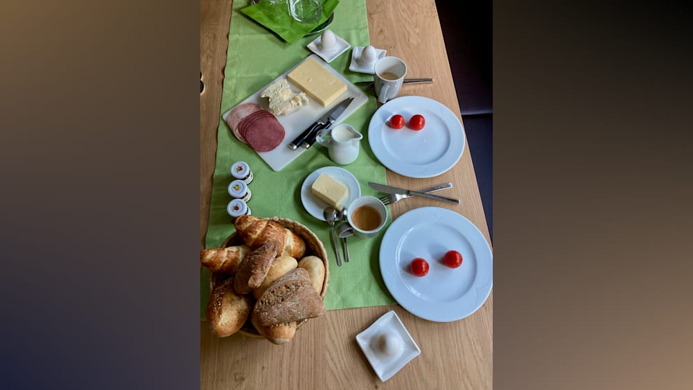 A table with plates, bread, butter, and tomatoes is set for two people to enjoy breakfast. One plate has a croissant and butter, while the other has a croissant and jam. There is also a basket of bread on the table.