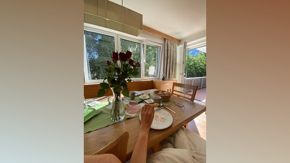 A person is sitting at a messy dining table with food and flowers.