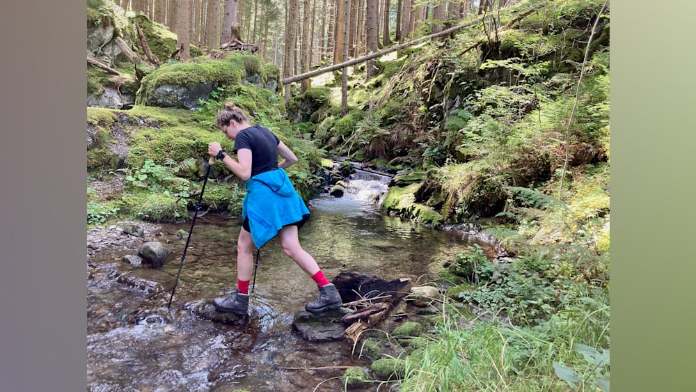 A woman is hiking and standing over water with a stick in her hand.