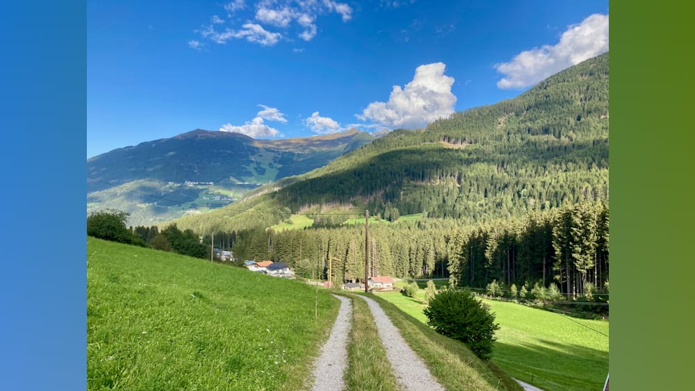 A road winds through a lush green valley with trees and houses in the background.