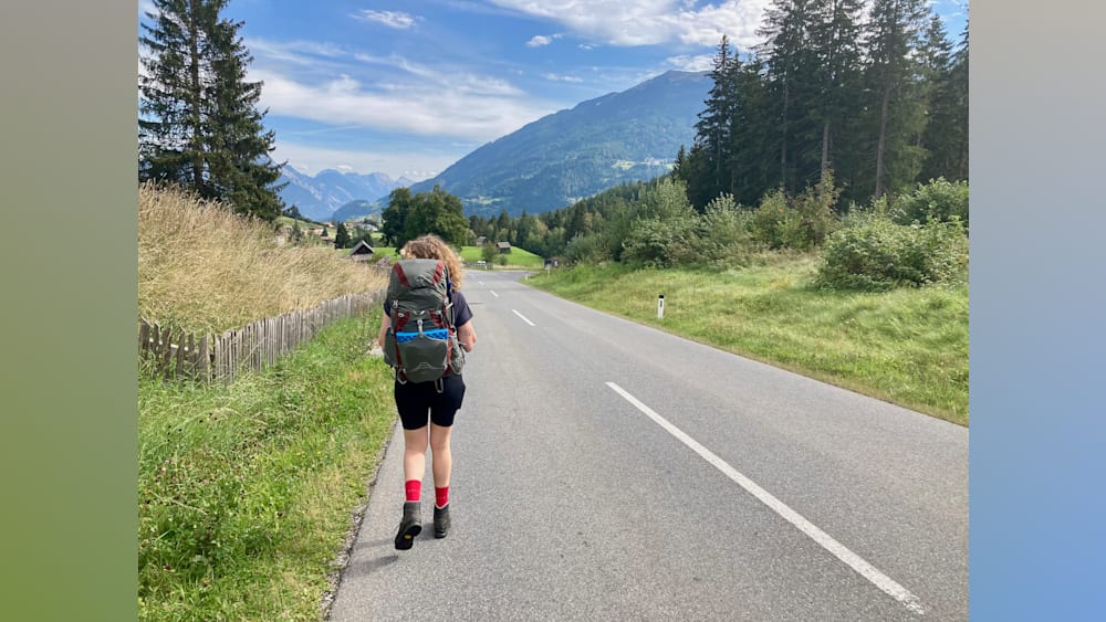 A woman wearing a black shirt is walking down a road with a grey and red backpack on her.