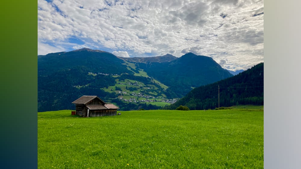 A cabin in the woods sits on a grassy field with mountains in the background.