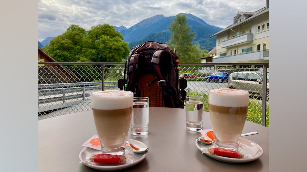 Two cups of coffee sit on a table with spoons and a backpack in the background.