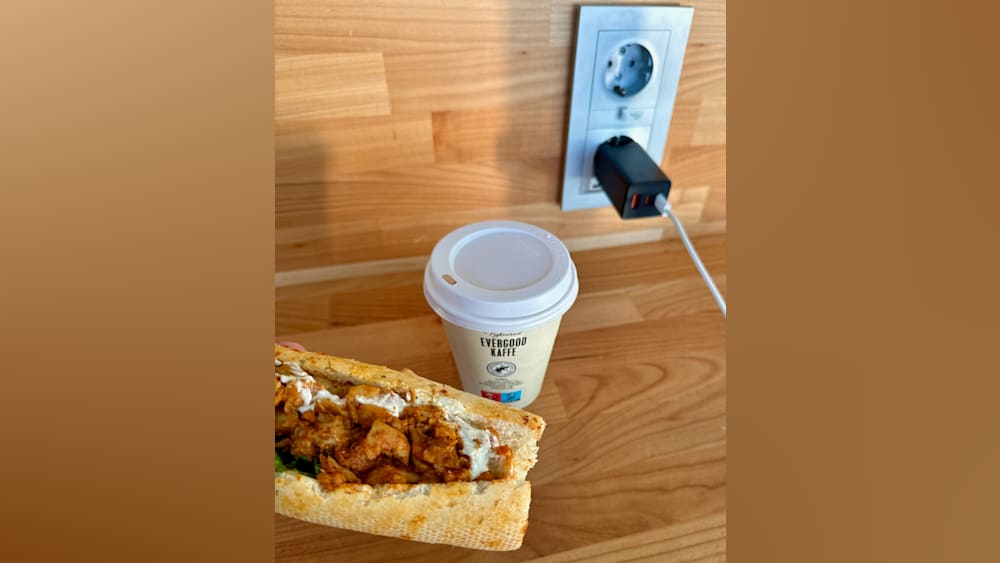 A cup of coffee next to a sandwich on a table with a plug in the wall.
