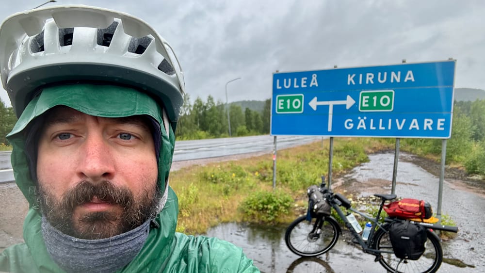A man wearing a green coat and helmet takes a picture in the rain in front of a blue sign with white arrow pointing right.